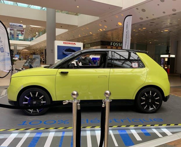 Gelber wasserstoffbetriebenes Elektroauto vor einem Gebäude mit Absperrpfosten, Bannern, Menschen, Schildern, einer Topfpflanze, Deckenleuchten und Ventilatoren.