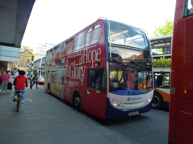 Eine Person fährt mit dem Fahrrad auf einem Bürgersteig mit vielen Menschen, Bussen mit Text darauf, Gebäuden und Laternen im Hintergrund.