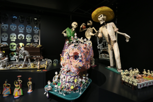 Eine Totenkopfausstellung im Fine Arts Museum in San Francisco, die Spielzeuge, Skelette und andere Gegenstände auf Tischen zeigt, mit einer Texttafel im Hintergrund, in einer dunklen und mysteriösen Atmosphäre.