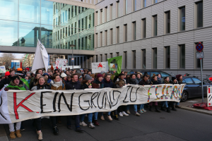 Eine Gruppe von Menschen steht auf einer Straße und hält ein Transparent mit der Aufschrift "Kein Grind Lo Feuer" hoch, im Hintergrund sind Gebäude, Bäume, Pfosten, Schilder, Fahrzeuge und ein klarer blauer Himmel zu sehen.