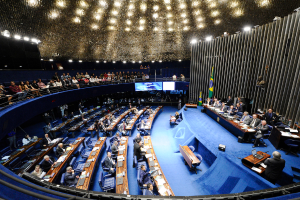 Eine Konferenzbühne mit sitzenden Personen an einem Tisch mit Papieren und Gegenständen, flankiert von Flaggen und einer Skulptur, mit Zuschauern an Tischen darunter und Beobachtern hinter einem Geländer mit Bildschirmen, unter dekorativen Deckenleuchten.