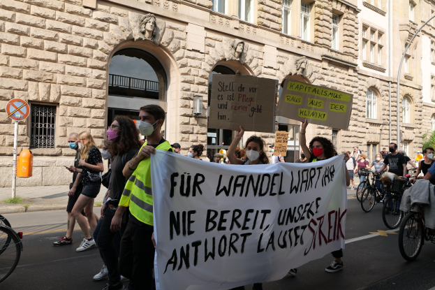 Eine Gruppe von Menschen marschiert durch eine Straße, einige halten Protestschilder und andere fahren Fahrráder, mit einem historischen Gebäude mit gewölbigen Fenstern, Pfeilern und Wandskulpturen im Hintergrund.