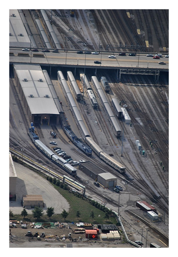 Eine Brücke mit wenigen Fahrzeugen darauf, darunter Eisenbahnschienen mit ein paar Zügen und eine Landschaft mit Gebäuden, Bäumen, Pfählen und diversen Objekten und Fahrzeugen am Boden.