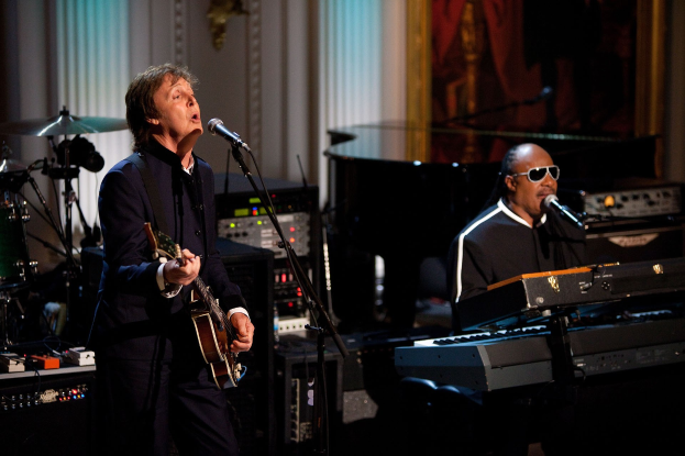 Paul McCartney spielt Gitarre und singt in ein Mikrofon, während Stevie Wonder am Piano sitzt und eine Brille trägt, bei der Grammy-Verleihung mit Musikinstrumenten und einem gerahmten Bild im Hintergrund.