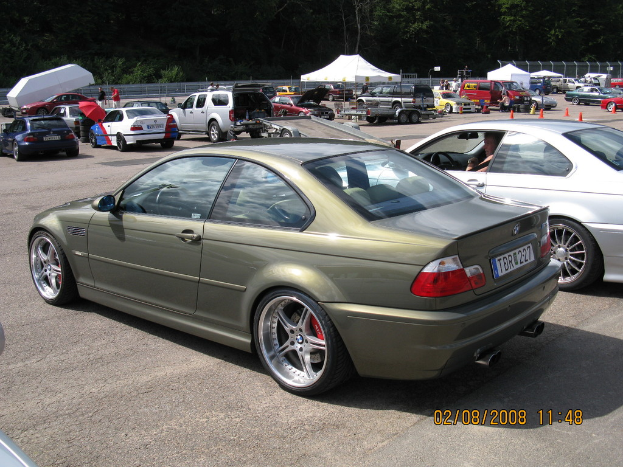 Eine Außenaufnahme mit mehreren Fahrzeugen auf einem Parkplatz, einem weißen Zelt in der Ferne, zahlreichen Bäumen, einigen stehenden Personen, einem roten Schirm und zwei Autos im Vordergrund.