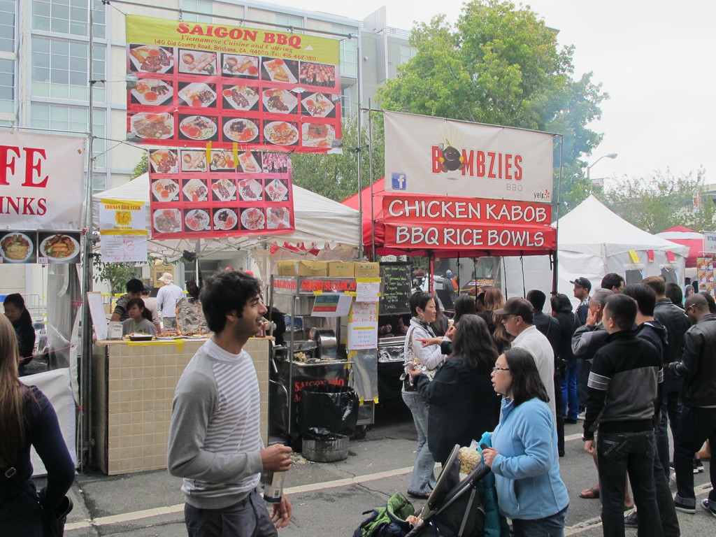 Menschen auf einer Straße vor der Stadt, mit einem Mann in einem grauen und weißen T-Shirt, der eine Wasserflasche hält, und einer Frau in einer blauen Jacke, die einen Kinderwagen schiebt, Stände mit weißen und roten Zelten, farbige Banner und Gebäude, Bäume und eine Straßenlaterne im Hintergrund.
