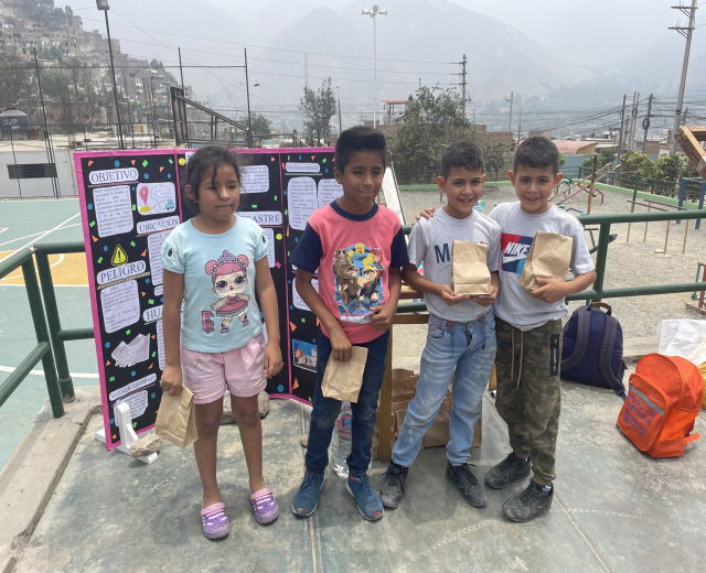 Gruppe von Kindern auf einem Basketballfeld mit Papiertüten, Vorräten auf dem Boden, einer Tafel mit Text links und einem eingezäunten Außenbereich mit Gebäuden und Hügeln im Hintergrund.