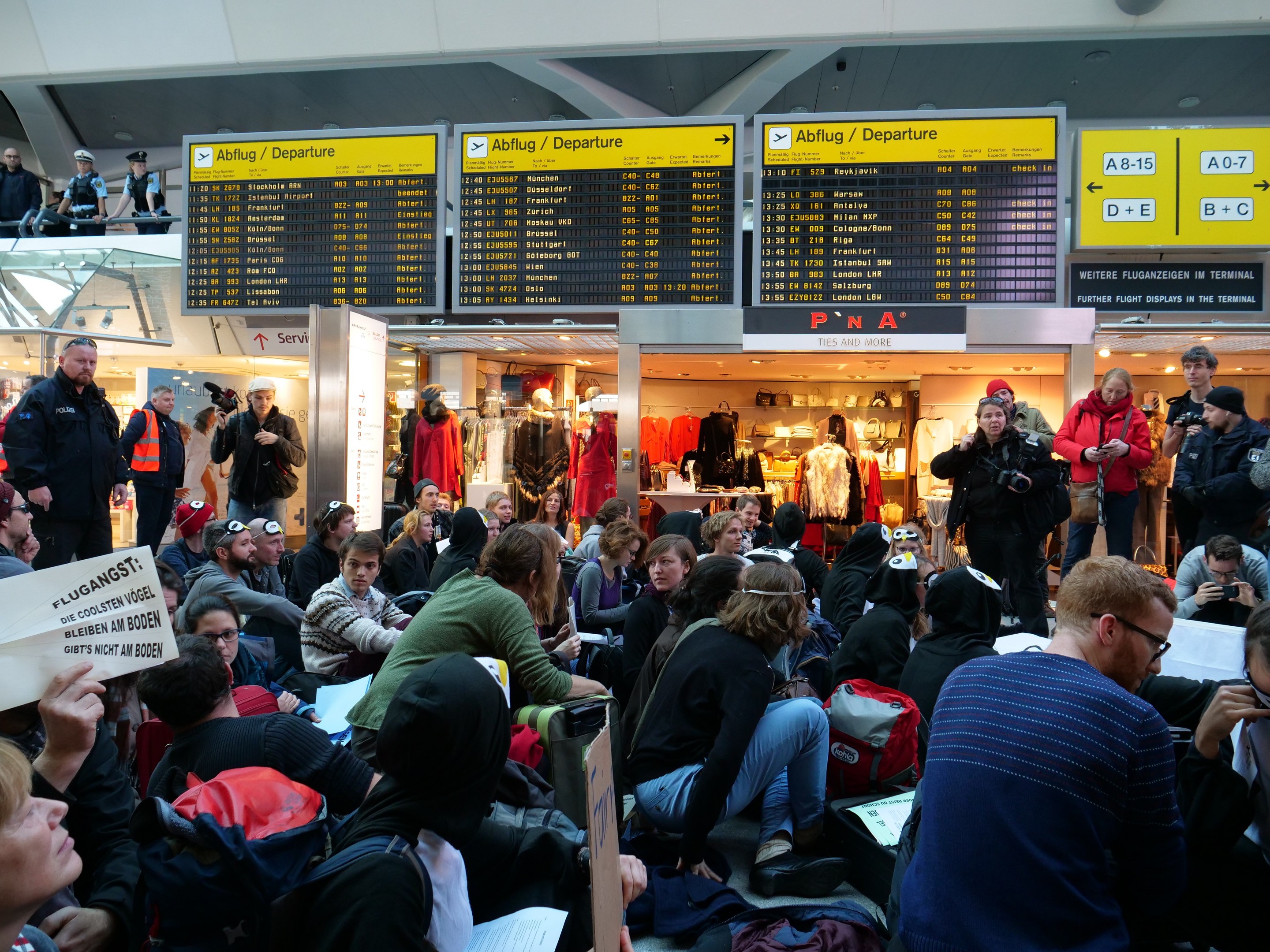 Große Gruppe von Menschen in einem Flughafen-Terminal, einige sitzen mit Taschen und Papieren, andere stehen, mit Informationsschildern, Schaufensterpuppen in Kleidern und Deckenbeleuchtung, was auf eine Protestaktion hindeutet.