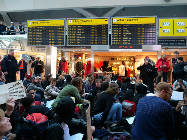 Große Gruppe von Menschen in einem Flughafen-Terminal, einige sitzen mit Taschen und Papieren, andere stehen, mit Informationsschildern, Schaufensterpuppen in Kleidern und Deckenbeleuchtung, was auf eine Protestaktion hindeutet.