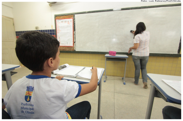 Ein Junge sitzt an einem Tisch und schreibt in einem Buch, während eine Frau die Tafel mit einem Putzlappen in einem Klassenzimmer mit mehreren Tischen, einem Plakat und einer Box auf einem Tisch abwischt.