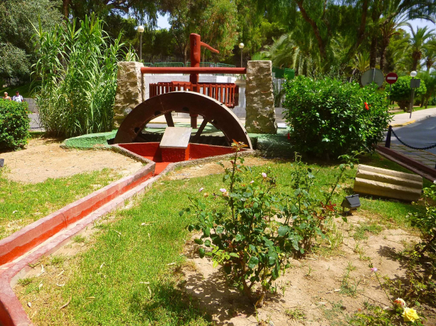 Ein Minigolfplatz in einem Park, umgeben von Grün, mit einer Straße, Passanten, Schildern und einem klaren blauen Himmel im Hintergrund.