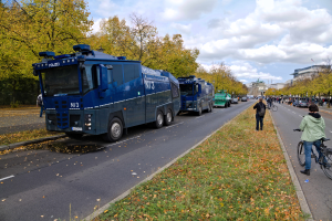 Eine Gruppe von Polizeifahrzeugen steht am Straßenrand geparkt, auf der rechten Seite fährt eine Person mit dem Fahrrad; viele Menschen, einige halten Fahrräder, stehen auf einem grasbewachsenen und blätterbedeckten Boden, im Hintergrund sind Bäume, Gebäude und ein bewölkter Himmel zu sehen.