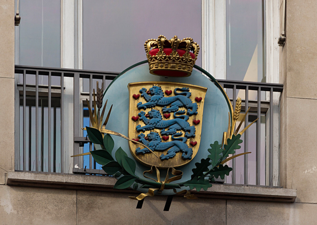 Wappen der britischen Königsfamilie auf der Fassade eines Gebäudes dargestellt, begleitet von einem Wappen mit Krone und Blättern, mit Fenstern und Metallgittern im Hintergrund.