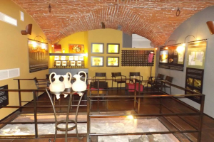 Ein Museumsraum voller Tische, Stühle, einem Geländer, Vasen, Texttafeln, Wandrahmen, Lampen und verschiedenen Ausstellungsstücken.