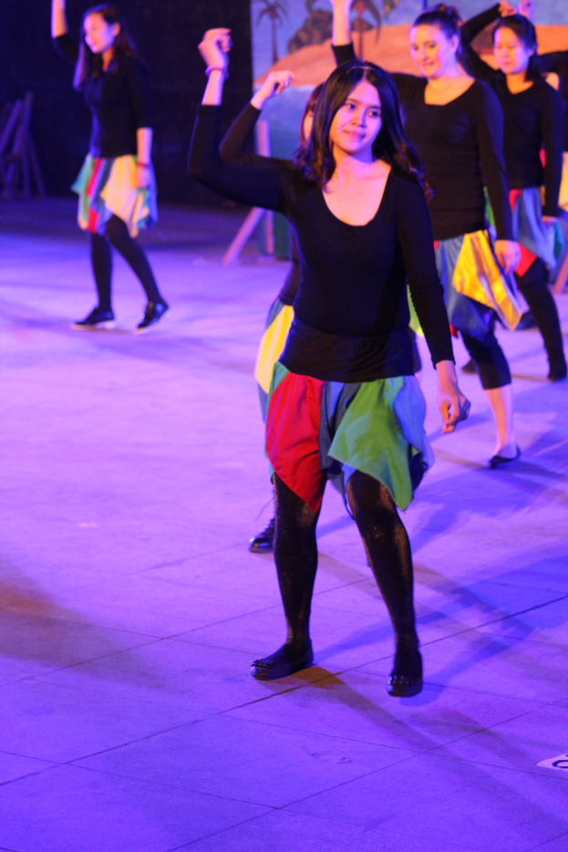 Gruppe junger Frauen, die auf einer Bühne tanzen, mit einem Gemälde im Hintergrund, alle tragen Schuhe während einer Vorstellung.