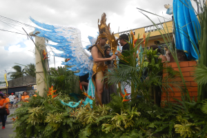 Eine Gruppe von Menschen, die eine Straße entlanggehen, neben einem Karnevalswagen mit Pflanzen und Blumen, Kostümierten, Gebäuden, Bäumen, Strommasten mit Drähten und einem bewölkten Himmel im Hintergrund.