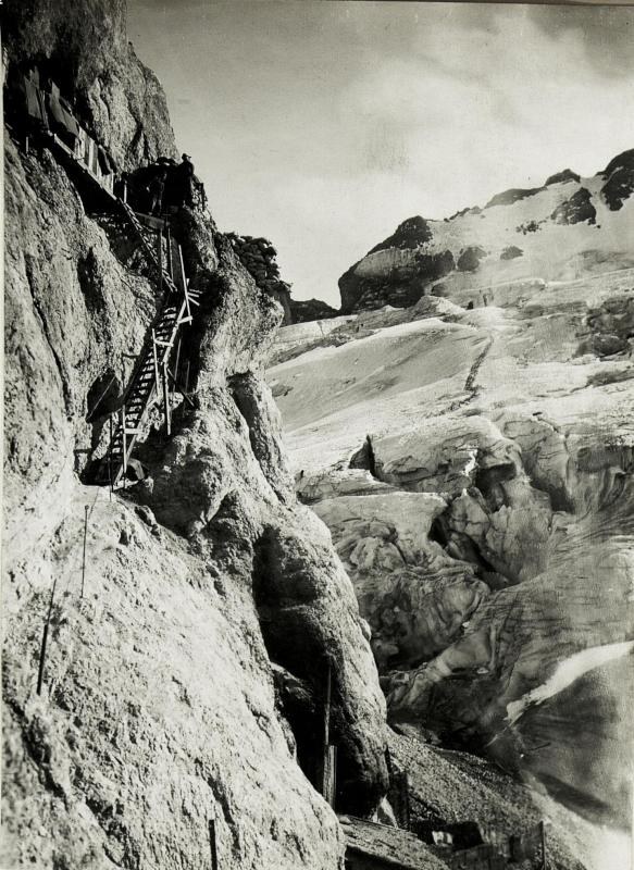 Schwarze und weiße Fotografie von Bergsteigern, die einen schneebedeckten Berg hinaufsteigen, mit fernen schneebedeckten Gipfeln und einem klaren Himmel, mit Text am unteren Rand.
