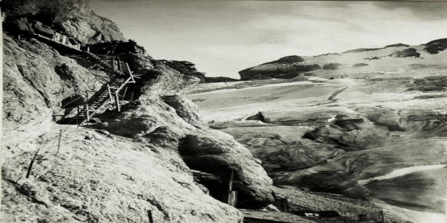 Schwarze und weiße Fotografie von Bergsteigern, die einen schneebedeckten Berg hinaufsteigen, mit fernen schneebedeckten Gipfeln und einem klaren Himmel, mit Text am unteren Rand.