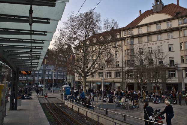 Eine belebte Stadtstraße mit einem Bahnhof auf der linken Seite, Menschen gehen, sitzen auf Bänken und stehen in der Nähe, umgeben von Gebäuden, Bäumen, Laternen und Schildern, mit dem Himmel im Hintergrund.