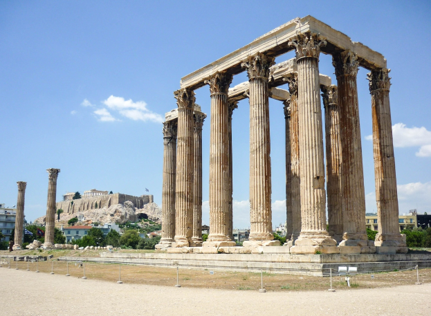 Alter Tempel des Olympian Zeus in Athen, Griechenland, mit hohen korinthischen Säulen, umgeben von Gebäuden, Bäumen und Felsen, mit einer Burg im wolkenverhangenen Himmel im Hintergrund.