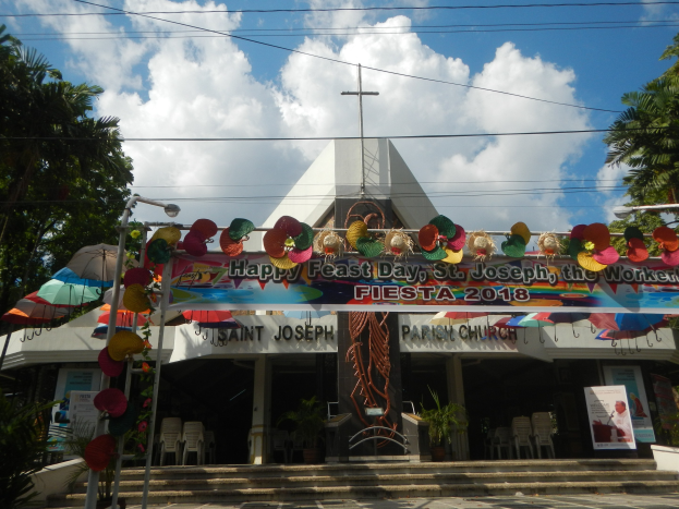 Ein Gebäude mit einer Tafel, die "Frohes Fest St. Josef der Arbeiter" trägt, davor, mit einer Treppe zum Eingang, einem Bogen mit hängenden Dekorationen, Außenbestuhlung mit Stühlen und Tischen, Topfpflanzen, Bäumen auf beiden Seiten und einem bewölkten Himmel.