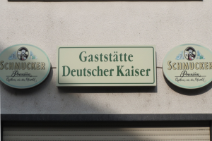 Drei hölzerne Bretter mit der Aufschrift "Gaststätte Deutscher Kaiser Schmucker" sind an einer Gebäudewand über einer geschlossenen Rolllade angebracht.