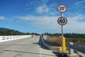 Ein Tempolimit-Schild am Straßenrand mit Motorrädern, Geländern, Bäumen, Bergen und einem klaren blauen Himmel im Hintergrund.