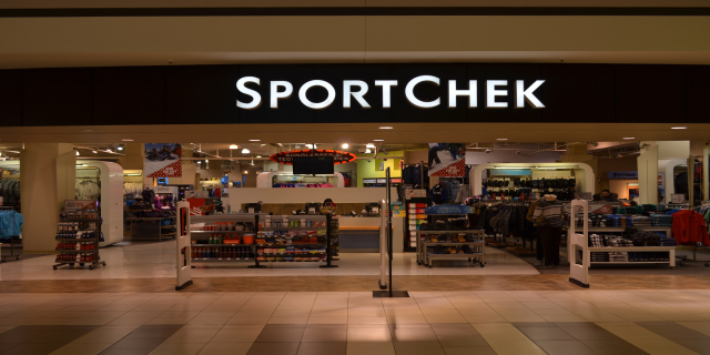 Außen- und Innenansicht eines Sportchek-Ladens in einem Einkaufszentrum mit Kleiderstangen, Deckenleuchten, Säulen und einem Fernseher.