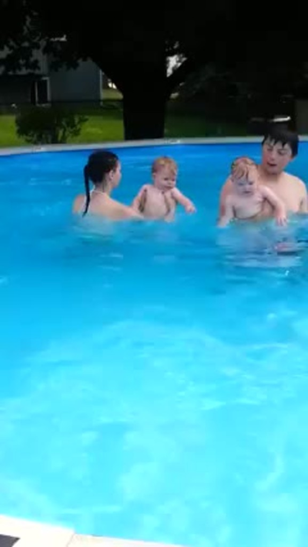 Ein Mann und eine Frau schwimmen in einem Pool, jeder hält ein Kind, mit Bäumen, Pflanzen und einem Gebäude im Hintergrund.
