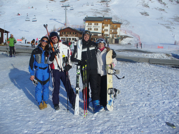 Vier Personen posieren für ein Foto mit Skating-Pads und Werkzeugen, mit Gebäuden und anderen Menschen im Hintergrund, vor einer schneebedeckten und vereisten Berglandschaft.