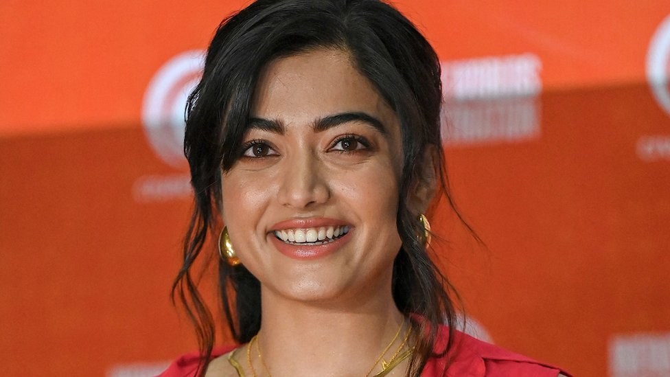 Eine Frau in einem roten Kleid mit einem eleganten Dutt und dezentem Make-up lächelt in die Kamera, trägt schwarze High Heels und hat ein hellgelbes Banner mit der Aufschrift "Sonam Kapoors beste Schönheits Looks vom Cannes Film Festival" im Hintergrund.