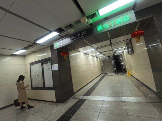 Eine Frau in einem cremefarbenen Kleid und einer schwarzen Tasche geht durch eine U-Bahn-Station mit einer Rolltreppe im Hintergrund, creme- und weißfarbene Wände und eine beleuchtete Decke.