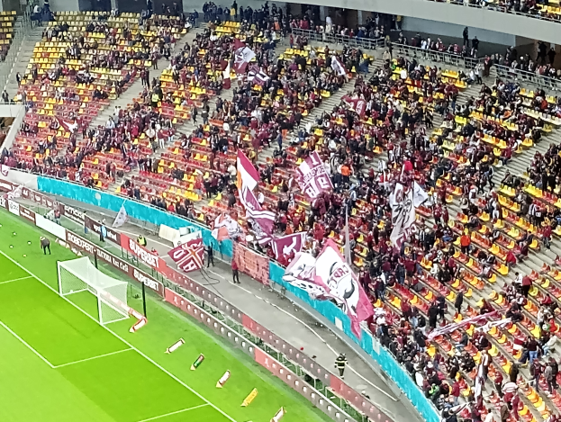 Ein großes Stadion voller Zuschauer bei einem Fußballspiel, mit einem Tor und Netz auf der linken Seite, Menschen, die stehen und sitzen, und Tafeln mit Text, Treppen, Geländer und eine Wand im Hintergrund.
