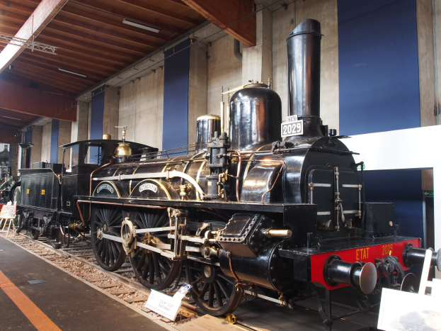 Eine große, alte Dampflokomotive in einem Museums Setting umgeben von Eisenbahnschienen, Informationsplakaten und anderen Ausstellungen, mit einer Wand und beleuchteter Decke im Hintergrund.
