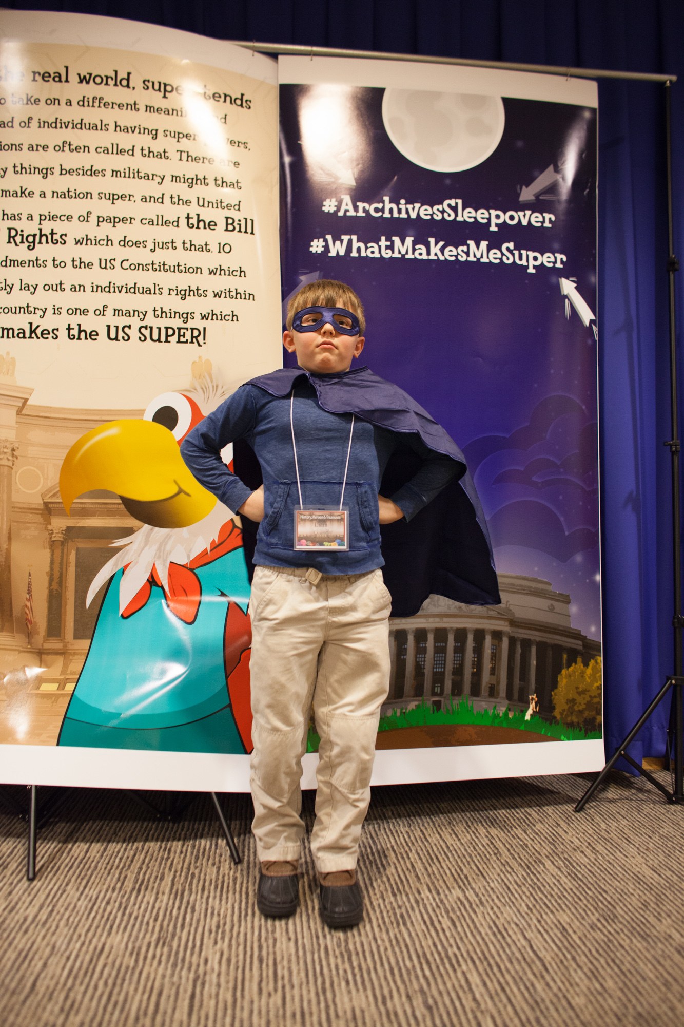 Ein junger Junge in einem Superheldenkostüm steht vor einem Banner mit Text und einem Vogelbild, mit einem Vorhang im Hintergrund und Schuhen.