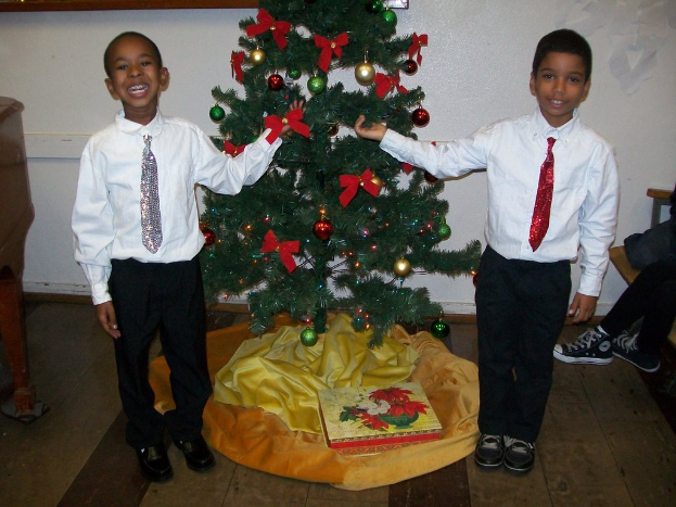 Ein geschmückter Weihnachtsbaum steht in einem Raum mit einem Holztisch links und einem Stuhl rechts, zwei Jungen in weißen Hemden lächeln und halten den Baum, eine Schachtel und andere Gegenstände liegen auf dem Boden, und eine weiße Wand mit angebrachten Gegenständen im Hintergrund.