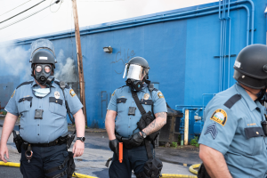 Drei Polizisten in Einsatzausrüstung, darunter Helme und Gasmasken, gehen eine Straße mit einer blauen Wand, Rohren und einem Strommast mit Kabeln im Hintergrund entlang, unter einem sichtbaren Himmel.