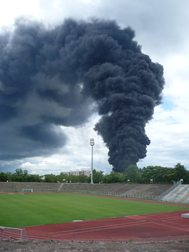 Eine große schwarze Rauchwolke steigt von der Spitze eines Stadions mit grasbewachsenem Boden, Stufen, Geländern, Bäumen, einem Lichtmast und einem bewölkten Himmel im Hintergrund auf.