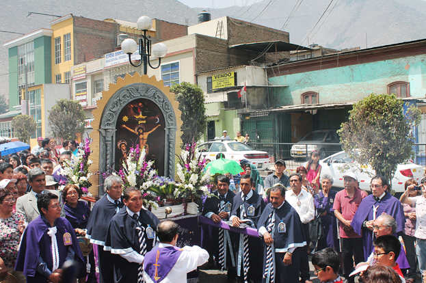 Menschen stehen auf einer Straße mit einem Kreuz und Blumensträußen in der Mitte, Autos fahren im Hintergrund und Gebäude, Bäume und Straßenlaternen sind weiter hinten zu sehen.