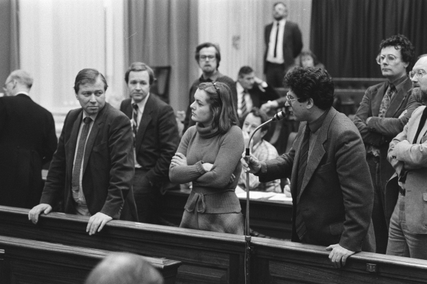 Ein Schwarz-Weiß-Bild einer Gruppe von Menschen in einem Gerichtssaal, mit einem Mann, der ein Mikrofon hält, Bänken im Vordergrund und Vorhängen und einer Wand im Hintergrund.