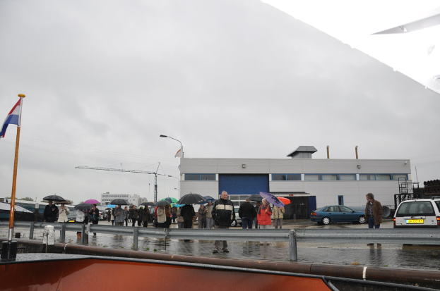 Eine Gruppe von Menschen im Freien, einige mit Schirmen, eine Fahne an einem Mast, zwei Autos, ein Gebäude mit Fenstern, ein Turmkran, eine Straßenlaterne und eine Absperrung.