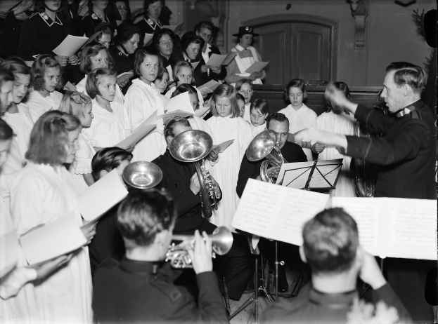 Eine Gruppe von Menschen steht vor einem Chor, jeder hält ein Buch und spielt verschiedene Musikinstrumente, dargestellt in Schwarz-Weiß.