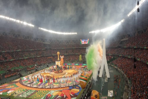 Ein großes Stadion mit Zuschauern bei einem Fussballspiel, das eine zentrale Bühne mit Menschen, Fahnen und Gegenständen zeigt, umgeben von sitzenden Gruppen auf beiden Seiten, mit Lichtern, einem Bildschirm und Rauch oben.