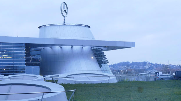 Das Mercedes-Benz Museum in Frankfurt, Deutschland, ein großes Gebäude mit einem Logo oben drauf, umgeben von Gras und Geländern, mit Fahrzeugen vorne auf der Straße und Bäumen unter einem klaren blauen Himmel im Hintergrund.