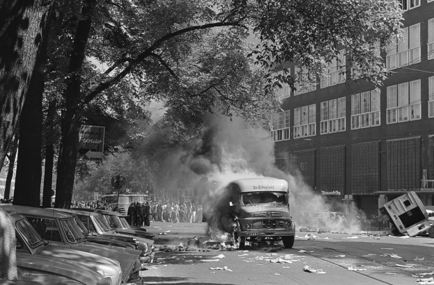 Schwarz-weiß-Foto eines brennenden Busses auf einer Straße mit parkenden Autos, Passanten, Bäumen, Gebäuden und einem Plakat im Hintergrund.