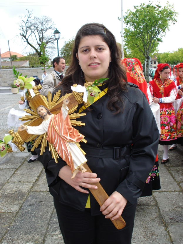 Eine Frau hält ein Abbild von Jesus Christus in der Hand, mit ein paar anderen Menschen, die dahinter auf dem Bürgersteig stehen, und Bäumen, Laternenpfählen, Pflanzen, Straßen, Gebäuden und einem Metallpfahl im Hintergrund.