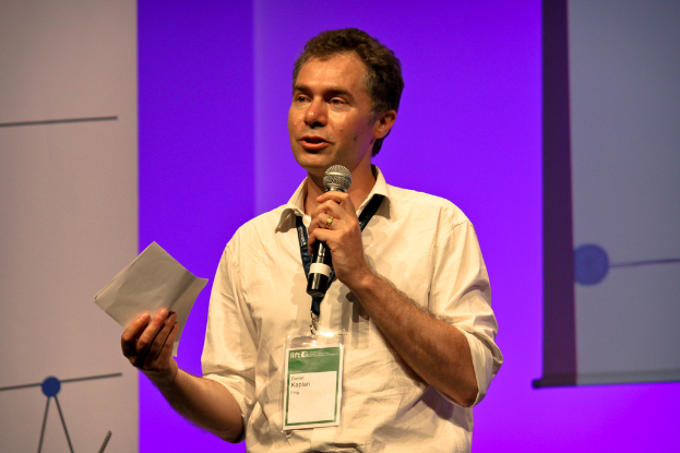 Ein Mann in einem cremefarbenen Hemd und Ausweis spricht in ein Mikrofon und hält ein Blatt Papier, vor einer lila Wand.