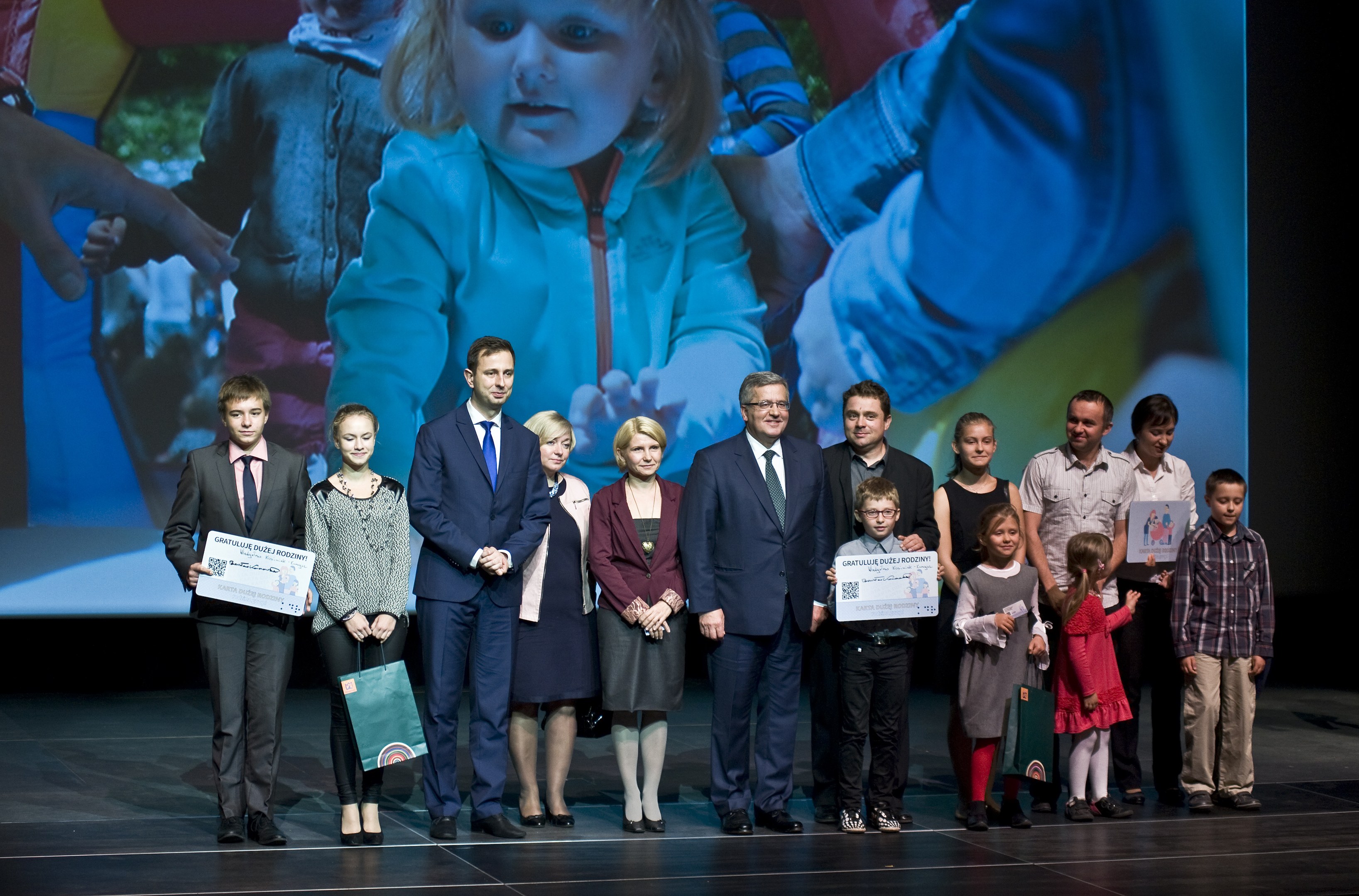Eine Gruppe von Menschen auf einer Bühne mit Taschen und Urkunden, mit einem Bildschirm, der ein Bild eines Kindes und andere im Hintergrund anzeigt.