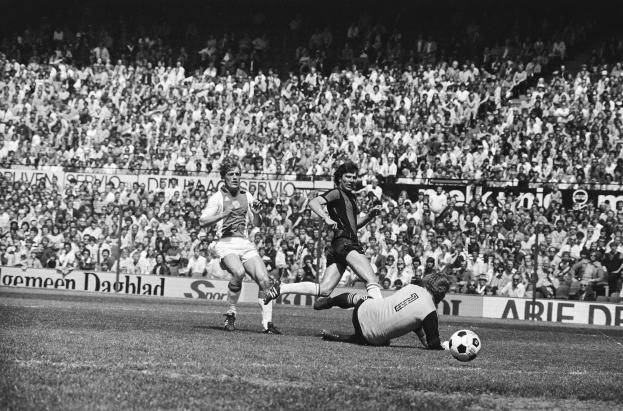 Schwarzes und weißes Foto von Männern, die auf einem Feld Fübball spielen, mit Zuschauern im Hintergrund und einer "Bundesliga 1978-79 Ajax 1-1 Hannover 96"-Tafel.
