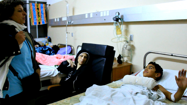 Ein Krankenhauszimmer mit mehreren Betten, auf denen Patienten liegen, Infusionsflaschen auf Ständern zwischen den Betten und eine Frau mit einer Tasche in der Nähe eines der Patienten